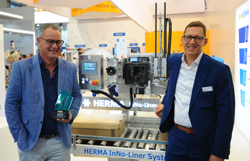 Zwei Männer stehen neben einem HERMA InNo-Liner System, einer Maschinenanlage zur Etikettierung, während einer von ihnen einen Preis hält.
