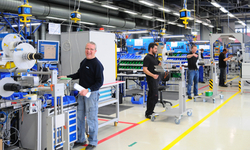 Arbeiter in einer Produktionshalle, die an Maschinen und Arbeitsplätzen stehen, während sie verschiedene Aufgaben ausführen.