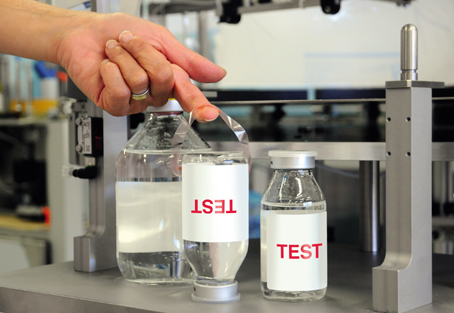 Eine Hand hebt ein Etikett mit der Aufschrift "TEST" von einer Flasche Wasser, während eine weitere Flasche daneben steht.