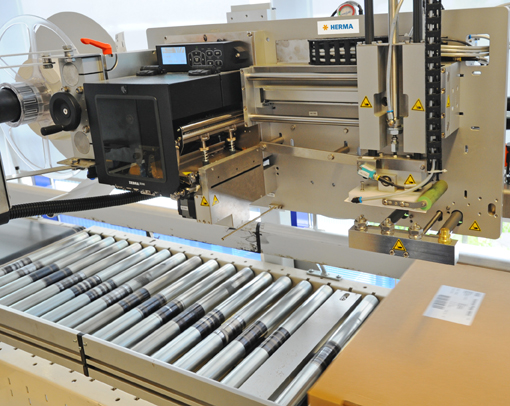 Eine Etikettiermaschine mit Rollenförderer, die Etiketten auf Pakete aufbringt. Die Maschine zeigt digitale Steuerungselemente und Sicherheitswarnungen.