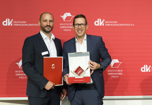 Zwei Männer stehen nebeneinander und halten Auszeichnungen in den Händen. Im Hintergrund ist ein rotes Banner mit dem Logo des Deutschen Verpackungsinstituts zu sehen.