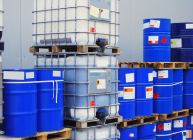 Gestapelte blaue Fässer und Container auf Paletten, gekennzeichnet mit verschiedenen Etiketten und Warnhinweisen.