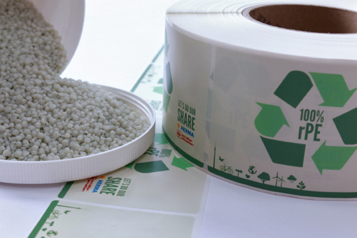 Grüne Kunststoffpellets liegen in einer offenen Dose, daneben eine Rolle mit umweltfreundlichem Etikett, das Recycling-Symbole zeigt.