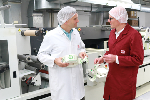 Zwei Personen in Schutzkleidung stehen in einer Produktionsumgebung und diskutieren über umweltfreundliche Verpackungsbanderolen.