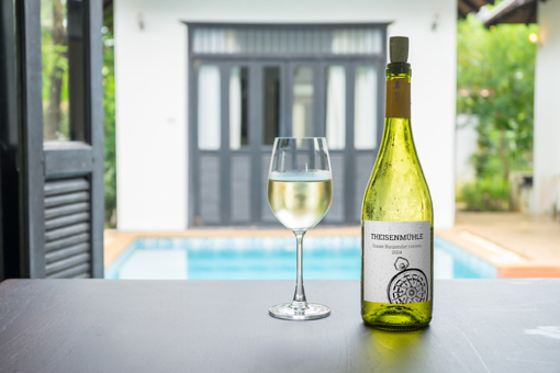 Eine Flasche Weißwein mit einem Glas auf einem Tisch, im Hintergrund ein Pool und ein helles, modernes Zimmer.