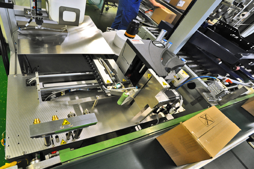 Automatisierte Verpackungsmaschine mit Förderbändern und Kartons im Hintergrund. Verschiedene mechanische Komponenten sind sichtbar.
