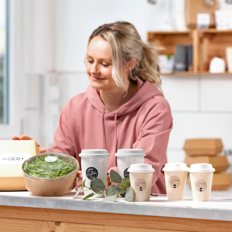 Eine Frau in einem rosa Hoodie bereitet gesunde Speisen vor, darunter eine Schüssel mit Salat und mehrere Kaffeebecher auf einem Tisch.