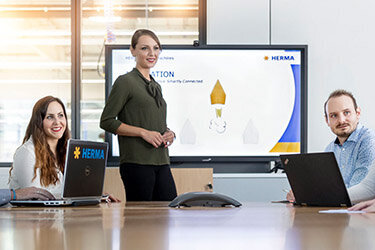 Eine Gruppe von sechs Personen sitzt an einem Konferenztisch, während eine Frau eine Präsentation vor einem Bildschirm hält.