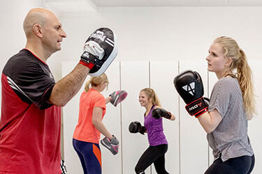 Eine Gruppe von Personen trainiert Boxen in einem hellen Raum. Zwei Teilnehmer üben mit Boxhandschuhen und Pratzen, während andere zuschauen.