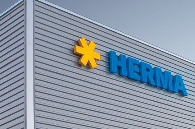 Fassade eines modernen Gebäudes mit dem Logo "HERMA" in blauer Schrift und einem gelben Symbol.