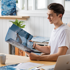Ein junger Mann sitzt an einem Tisch und betrachtet einen Ordner mit Bildern von Wellen und Meer. Ein Laptop und eine Tasse sind ebenfalls sichtbar.