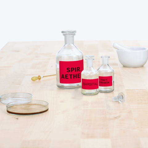Flaschen mit roten Etiketten, beschriftet mit "Spir. Aether", "Barbital" und "Tinct. Tramon", auf einem Holztisch. Ein Mörser und zwei Deckel sind ebenfalls sichtbar.