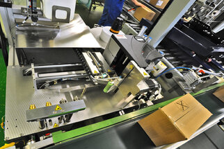 Automatisierte Verpackungsmaschine mit Förderbändern und Kartons im Hintergrund. Verschiedene mechanische Komponenten sind sichtbar.