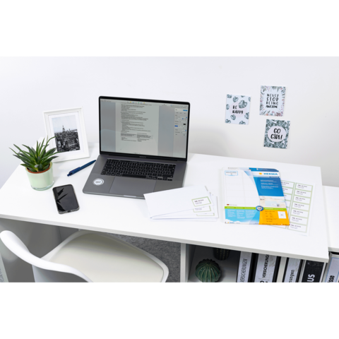 Ein Schreibtisch mit einem Laptop, einem Smartphone, einer Pflanze, Briefumschlägen und Dokumenten sowie dekorativen Wandbildern.