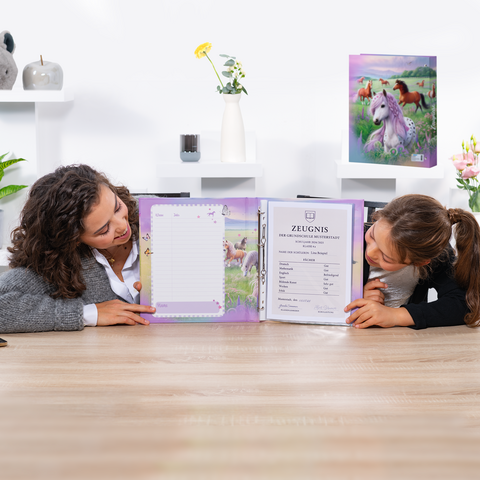 Zwei Mädchen sitzen am Tisch und betrachten ein offenes Schulzeugnis in einem bunten Ordner mit Einhorn-Motiv.