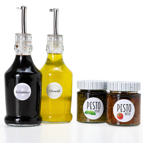 Zwei Flaschen mit Balsamico und Olivenöl sowie zwei Gläser Pesto in verschiedenen Sorten auf einem weißen Hintergrund.