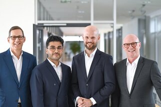 Vier Männer in Anzügen stehen in einem modernen Bürogebäude. Sie lächeln und posieren freundlich für die Kamera.