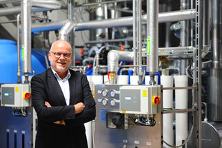 Ein Mann in einem Anzug steht lächelnd vor einer industriellen Wasseraufbereitungsanlage mit Rohren und Steuergeräten.
