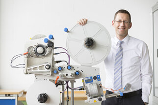 Ein Mann in einem Hemd und einer Krawatte steht neben einer technischen Maschine mit Rollen und Steuerungselementen.