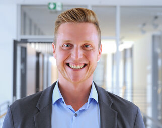 Ein lächelnder Mann mit blonden Haaren trägt einen grauen Anzug und ein hellblaues Hemd. Er steht in einem modernen Innenraum.