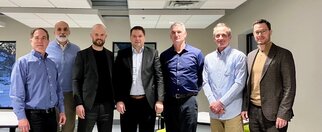 Eine Gruppe von sieben Männern steht in einem modernen Raum, alle in formeller Kleidung, mit verschiedenen Hemden und Anzügen.