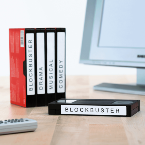 Eine Sammlung von Videokassetten mit der Aufschrift "BLOCKBUSTER", darunter Genres wie Drama, Musical und Comedy, neben einem Computer.