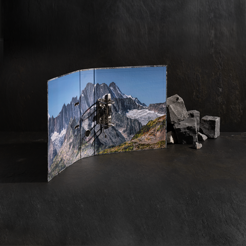 Eine aufgeschlagene Buchseite zeigt eine Berglandschaft mit einem alten Gerät, daneben liegen einige Steine.