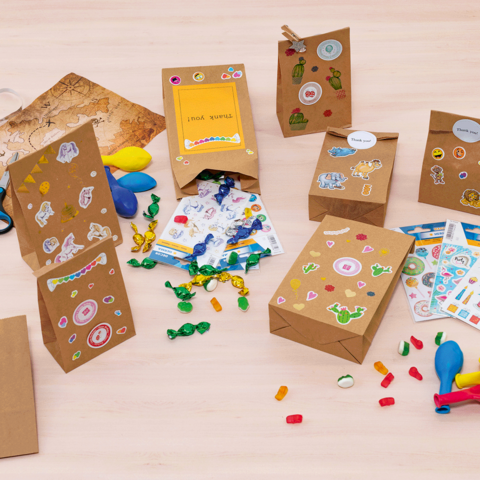 Bunte Geschenktüten und Bastelmaterialien auf einem Tisch, darunter Aufkleber, Luftballons, Scheren und Süßigkeiten.
