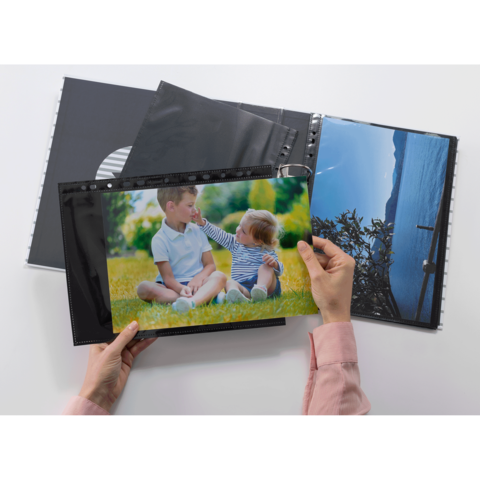 Eine Hand hält ein Foto von zwei Kindern, die im Freien auf dem Gras sitzen und miteinander interagieren, während ein Fotoalbum geöffnet ist.