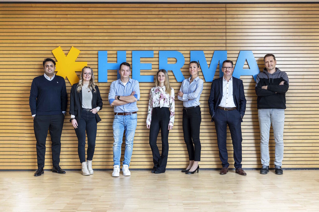 Eine Gruppe von sieben Personen steht vor einer Holzwand mit dem Schriftzug "HERMA". Sie posieren in formeller Kleidung.