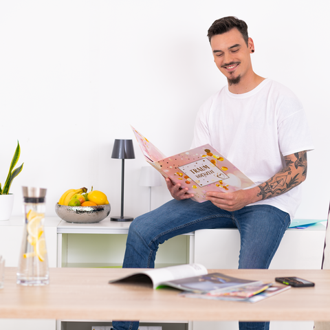Ein Mann mit Tätowierungen sitzt an einem Tisch und liest ein Buch. Auf dem Tisch stehen eine Wasserflasche und Obst.