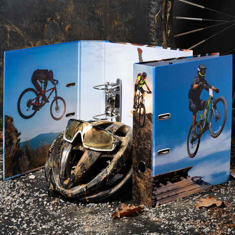 Ein schmutziger Fahrradhelm liegt vor einem offenen Ordner mit Bildern von Mountainbikern, die über hügelige Landschaften fahren.