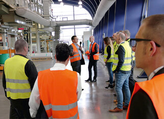 Eine Gruppe von Personen in Sicherheitswesten hört einem Referenten in einer Industrieumgebung zu.
