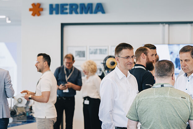 Menschen in formeller Kleidung stehen in einem modernen Raum, diskutieren und interagieren, mit einem HERMA-Schild im Hintergrund.
