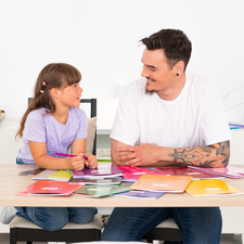 Ein Mann und ein Mädchen sitzen an einem Tisch, umgeben von Farbmustern und Materialien, während sie miteinander sprechen.