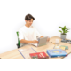 Ein junger Mann sitzt an einem Tisch, nutzt ein Tablet und hat mehrere Bücher sowie Schreibmaterialien vor sich. Eine Pflanze ist im Hintergrund.