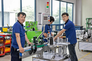 Drei Arbeiter in blauen Shirts bedienen eine Maschinenanlage in einer Fabrik, umgeben von Werkzeugen und Maschinen.