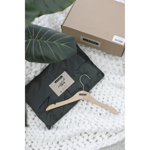 Ein schwarzes Paket mit einem Aufkleber und einem Holzkleiderbügel liegt auf einer weißen, gestrickten Decke, umgeben von einer Pflanze.