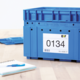 Blaue Kunststoffbox mit einer Etikette, die die Nummer 0134 und weitere Details zeigt, auf einem Tisch neben einem Notizblock und einem Stift.