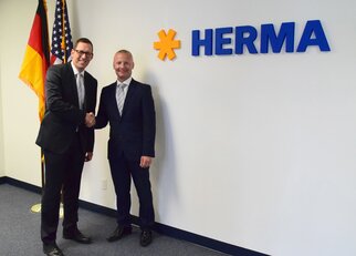 Zwei Männer in Anzügen schütteln sich die Hände vor einer Wand mit dem Schriftzug "HERMA" und Flaggen von Deutschland und den USA.