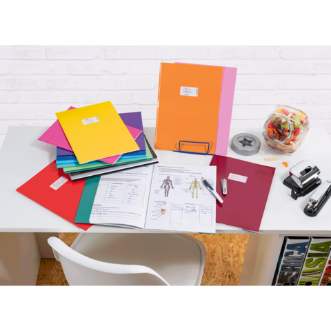 Bunte Ordner und Unterlagen auf einem Schreibtisch, daneben ein Glas mit Süßigkeiten und Bürobedarf wie einen Hefter und einen Stift.