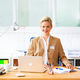 Eine Frau mit blonden Haaren sitzt an einem Schreibtisch, lächelt und trägt einen beigen Blazer. Vor ihr steht ein Laptop und Schreibmaterial.