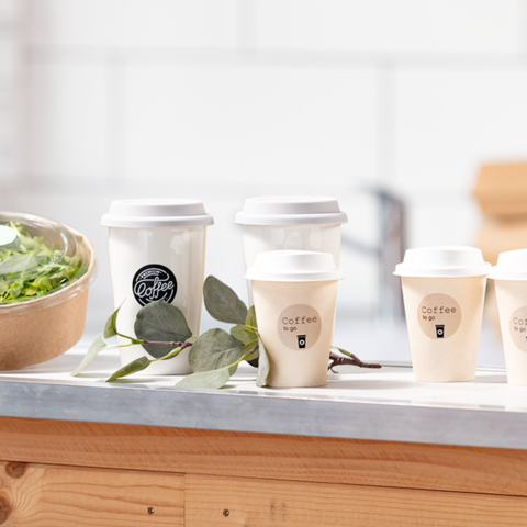Mehrere Kaffeebecher stehen auf einem Tisch neben einer Schüssel mit Salat. Die Becher sind mit Aufschriften zum Mitnehmen versehen.