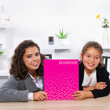 Zwei Mädchen sitzen an einem Tisch und halten ein pinkes Heft mit der Aufschrift "ZEUGNISSE". Sie lächeln in die Kamera.