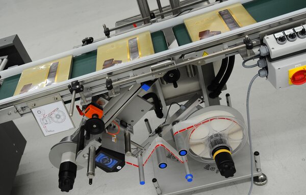 Ein Förderband mit verpackten Produkten, ausgestattet mit einer Etikettiermaschine und verschiedenen Steuerungselementen.