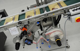 Ein Förderband mit verpackten Produkten, ausgestattet mit einer Etikettiermaschine und verschiedenen Steuerungselementen.