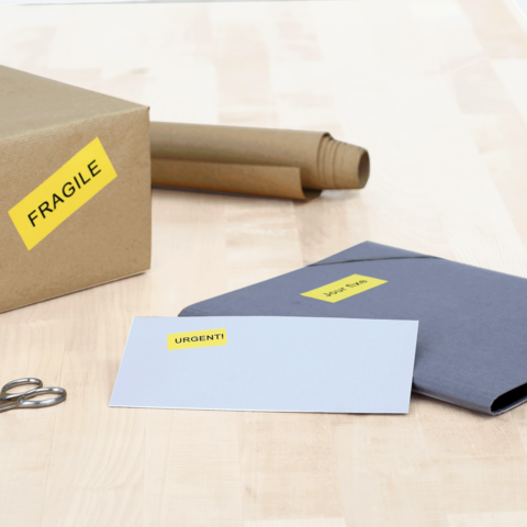 Ein brauner Karton mit dem Aufkleber "FRAGILE", daneben ein blauer Umschlag mit "DRINGEND!" und eine Schere auf einem Holztisch.