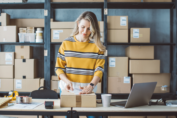 Eine Frau in einem gestreiften Pullover steht an einem Tisch, umgeben von Kartons und einem Laptop in einem modernen Büro.