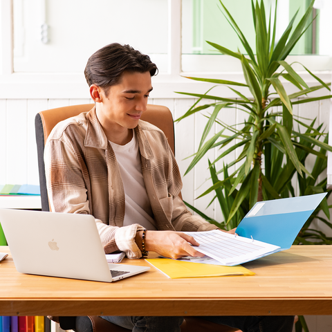 Ein junger Mann sitzt an einem Schreibtisch, arbeitet mit einem Laptop und blättert durch Unterlagen, umgeben von Pflanzen und bunten Ordnern.