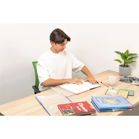 Ein junger Mann sitzt an einem Tisch, blättert in einem Notizbuch und hat Bücher sowie eine Pflanze und eine Tasse neben sich.